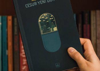 Crop unrecognizable person demonstrating book with illustration of contemporary buildings and author name on front cover near wooden bookshelf with assorted literature in house