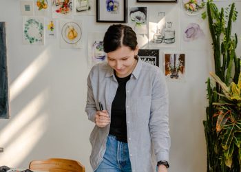 Cheerful young female designer in jeans standing near wall with pictures and table with stationery while choosing drafts with letter design in daylight