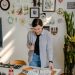 Cheerful young female designer in jeans standing near wall with pictures and table with stationery while choosing drafts with letter design in daylight