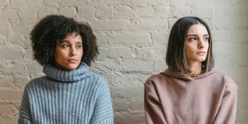 Sad African American woman with female friend in casual clothes looking away while sitting in light room during quarrel at home
