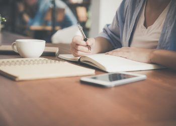 Woman Writing on a Notebook Beside Teacup and Tablet Computer